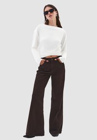 Bílá košile s dlouhým rukávem a řasením v pase, doplněná o tmavě hnědé rozšířené kalhoty, obě jsou pohodlně padnoucí. Nasazeny černé sluneční brýle.