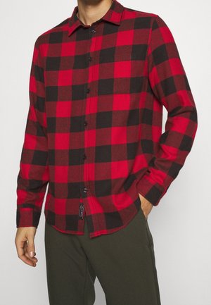 Mężczyzna w czerwono-czarnej koszuli w kratę na guziki z długimi rękawami, jedną ręką w kieszeni, w ciemnozielonych spodniach.
