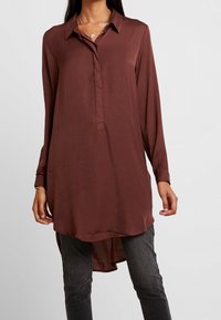 Femme portant une tunique marron à manches longues et à boutons sur un pantalon gris foncé, avec un collier et un bracelet en argent visibles.