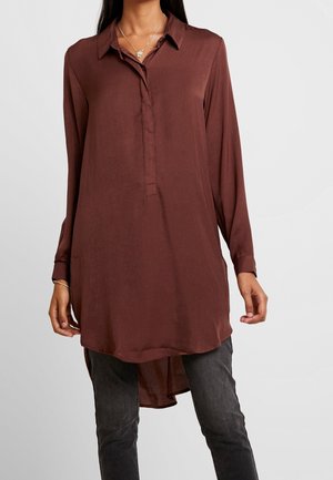 Kvinde iført en langærmet, knappet, brun tunika skjorte over mørkegrå bukser, med en sølvhalskæde og armbånd synligt.