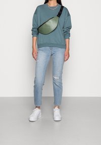 Sac bandoulière en cuir vert porté sur un sweat-shirt bleu, associé à un jean bleu clair déchiré et des baskets blanches. Chaîne de cou visible.