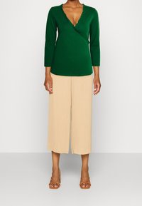 Grünes Langarm-Wickeloberteil mit gerafftem Ausschnitt, kombiniert mit beigen, plissierten Weitbein-Hosen und braunen Riemchensandalen.