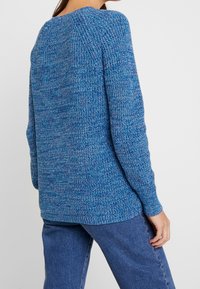 Blå stickad tröja med en texturerad mönster, rund halsringning och långa ärmar. Bärs med blå denimbyxor som har en avslappnad passform och drapering.