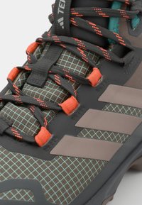 Grå og brun tur- og fjellsko med en teksturert overdel, turkis detaljer, oransje hardware og et mønstrete rutenett-design på materialet.