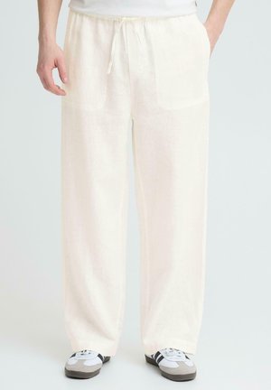 Person iført løse, cremefarvede bukser med snøre i taljen og forlommer samt hvide sneakers med sorte striber, stående mod en ensfarvet baggrund.