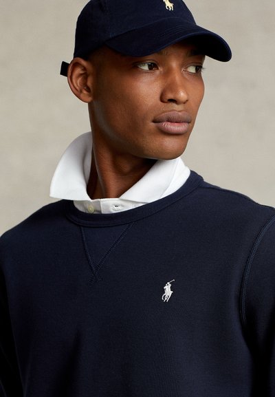Uomo che indossa un berretto blu navy e una felpa blu navy abbinata con il colletto bianco della polo in vista, guarda verso destra con un'espressione neutra.