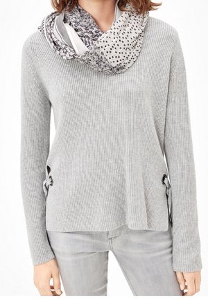 Femme portant un pull côtelé gris clair avec des liens sur les côtés, un jean gris et une écharpe infinity à motifs gris et blanc.