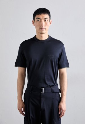 Mörk marinblå t-shirt med kort ärm och rund hals, försedd med en subtil logotyp på bröstet. Kombinerad med högmidjade byxor och ett silverbälte.