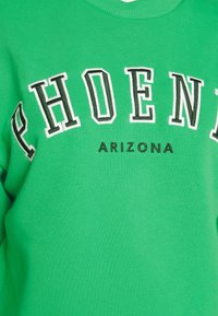 Grön sweatshirt med "PHOENIX" i fet stil i vita bokstäver med svarta konturer och "ARIZONA" i mindre svart text nedanför. Mjuk tygkänsla.