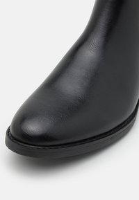 Botín negro con una superficie de cuero sintético suave, punta afilada y una suela negra texturizada de baja altura. Diseño sencillo sin herrajes visibles.