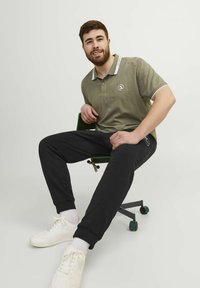 Žalias polo marškinėliai su baltais akcentais, juodos sportinės kelnės ir baltos sportbačiai. Modelis sėdi ant žalios kėdės, šypsodamasis.
