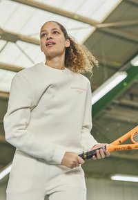 Felpa beige chiaro e pantaloni abbinati, con un logo in pesca, dalla texture liscia. Il soggetto tiene in mano una racchetta da tennis di un brillante arancione.