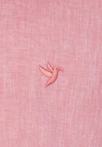 Tela de color rosa claro con un acabado texturizado que presenta un pequeño colibrí bordado en un hilo de rosa ligeramente más oscuro.