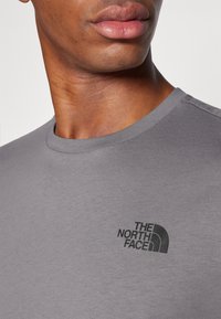 Pilkas marškinėlis, pagamintas iš švelnios medžiagos, su apvaliu kaklu ir juodu „The North Face“ logotipo spaudu kairėje krūtinės pusėje.