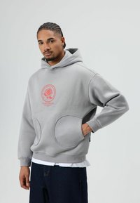 Grijze hoodie met grote ronde zakken en een rood rozenmotief op de borst. Gemaakt van zacht materiaal, met een relaxte pasvorm.