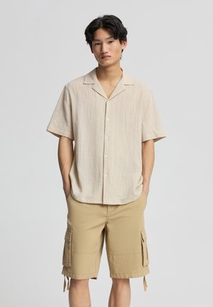 Giovane uomo con camicia beige a maniche corte abbottonata e pantaloncini cargo color sabbia, in piedi con le mani nelle tasche davanti a uno sfondo semplice.