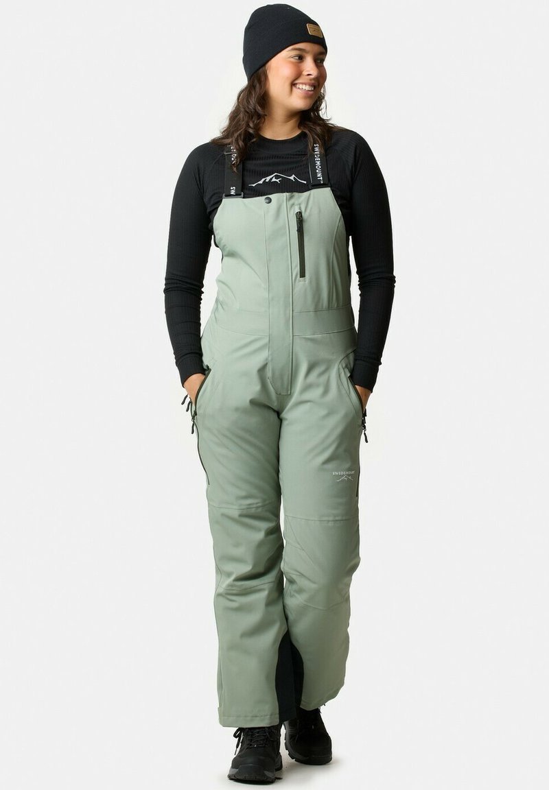 Ljusgröna isolerade hängselbyxor med justerbara axelband, ficka med dragkedja på bröstet och sidofickor, i kombination med en svart långärmad topp.