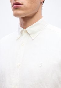 Gros plan d'un homme portant une chemise blanche à boutons avec un col boutonné et un logo brodé subtil sur la poitrine.