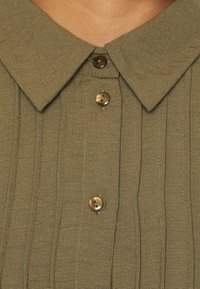 Olive green shirt with a collar, horizontal pleats, and two brown buttons on the front. Soft, textured fabric with a subtle weave pattern.
