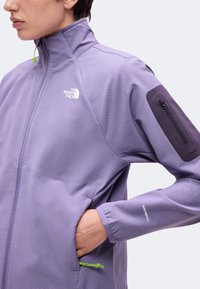 Άτομο που φοράει μωβ μπουφάν North Face με τσεπάκι με φερμουάρ στο μανίκι, χέρι στην πλαϊνή τσέπη και κείμενο "Windwall" στο μανίκι.