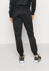 Personne portant un pantalon de jogging noir fuselé et des baskets noires à semelles blanches, debout de dos sur un fond uni.