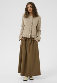 Mlada ženska z kodrastimi lasmi, oblečena v bež pulover z volančki in dolg rjav krilo ter kremo barvo čevljev, stoji z rokami v žepih.