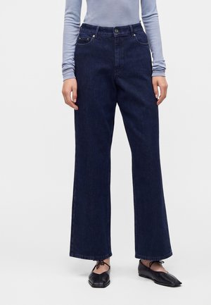 Person iført mørkeblå højtaljede vidde jeans, lyseblå langærmet top og sorte flade snørebåndssko, der står op ad en ensfarvet baggrund.