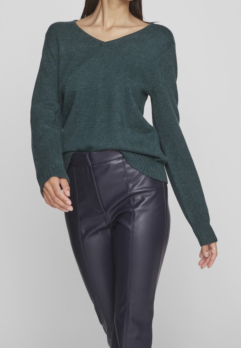 Femme portant un pull vert foncé à col en V et un pantalon en cuir violet foncé sur un fond clair uni.