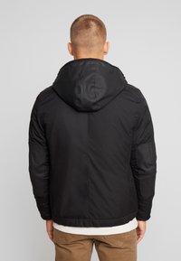 Black padded jacket with a hood and zippered pockets. Features ribbed cuffs and a smooth finish, showcasing a minimalist design.