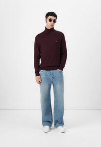 Pull en col roulé bordeaux, jean bleu délavé à coupe décontractée et baskets blanches. Le mannequin porte des lunettes de soleil ; marquage minimal sur le pull.
