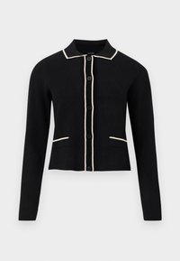 Melnā kardigāna ar apkakles dekoltē, krēmkrāsas apdari ap apkakli un kabatām, kā arī sešām priekšējām pogām. Tam ir garas piedurknes un īsa piegriezte.