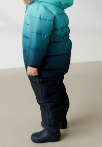 Mono de invierno acolchado en degradado de azul verdoso a negro, con capucha, cinturilla elástica y botas de goma negras con suelas texturizadas.