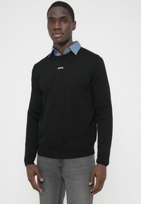 Pulover negru tricotat cu guler rotund, având un mic logo alb „HUGO” pe piept, purtat peste o cămașă bleu deschis cu guler.