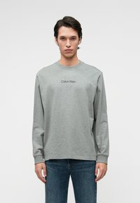 Γκρι μακρυμάνικη μπλούζα με στρογγυλή λαιμόκοψη και διακριτό λογότυπο Calvin Klein στο κέντρο του στήθους. Καθημερινή εφαρμογή, συνθετικό βαμβακερό υλικό.