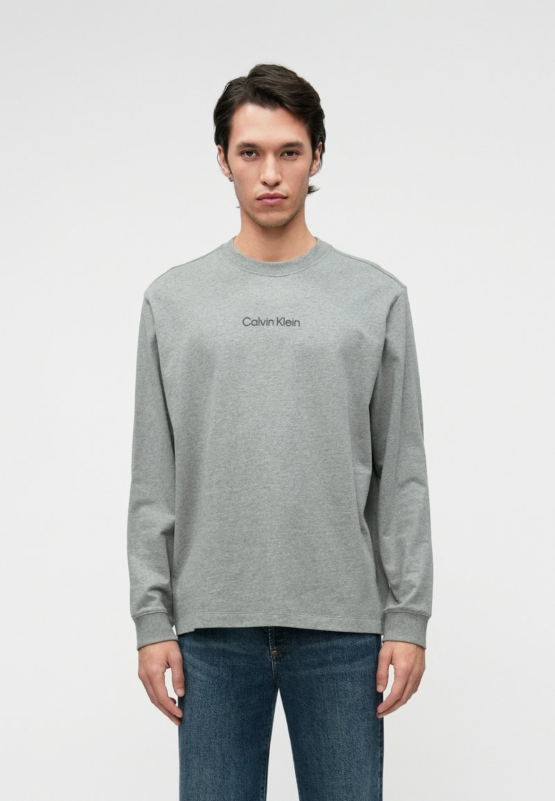 Γκρι μακρυμάνικη μπλούζα με στρογγυλή λαιμόκοψη και διακριτό λογότυπο Calvin Klein στο κέντρο του στήθους. Καθημερινή εφαρμογή, συνθετικό βαμβακερό υλικό.