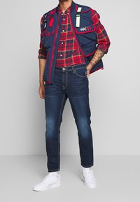 Mörkblå vadderad väst med röda detaljer över en röd och grön rutig skjorta, i kombination med mörk denimjeans och vita sneakers.