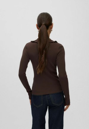 Femme aux longs cheveux bruns attachés en queue de cheval, portant un haut marron moulant à côtes à manches longues et un jean bleu foncé, debout de dos sur un fond uni.