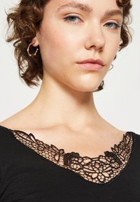 Svart topp med en intrikat svart blondehalsutskjæring med blomsterdesign, som fremhever tekstur og detaljerte mønstre.