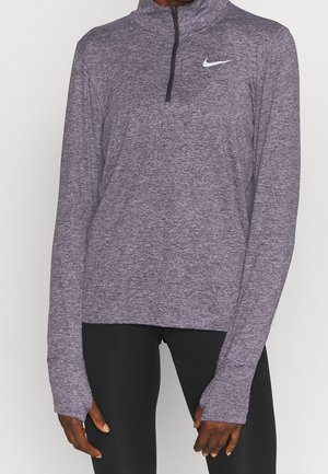 Person trägt ein violettes langärmeliges Nike-Sporttop mit Daumenlöchern und schwarze Leggings, steht vor einem einfarbigen Hintergrund.