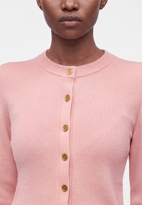 Femme portant un cardigan côtelé rose avec cinq boutons dorés visibles, montrée des épaules au menton sur un fond uni.