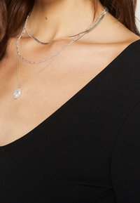 Tre lager av silverhalsband, med en texturerad berlock. Bärs över en svart topp. Delikata kedjedesigner och varierande längder.