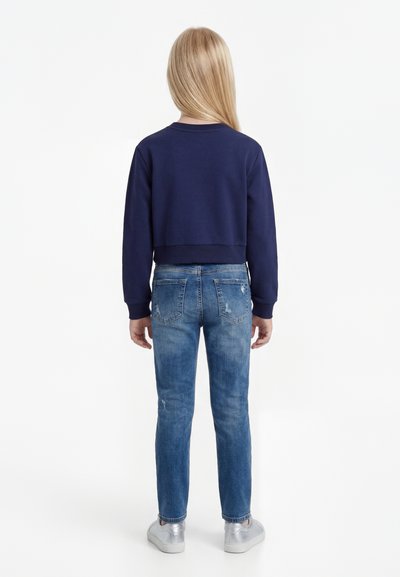 Hosszú szőke hajú lány tengerészkék pulóvert, kék farmert és ezüst tornacipőt visel, háttal áll fehér háttér előtt.