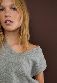 Sivi pulover bez ramena sa širokim izrezom i rebrastim rubovima. Mekani materijal, labav kroj i kratke rukave na tamnosmeđoj pozadini.