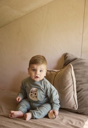 Bambino piccolo con abito verde con stampa di cane, seduto a piedi nudi su un letto beige, tiene un blocco di legno, una parete di legno neutra e cuscini sullo sfondo.