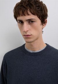 Granatowy sweter z okrągłym dekoltem wykonany z miękkiego dzianego materiału, noszony na jasnoszarej koszulce, z prążkowanym wykończeniem na szyi i delikatną fakturą.