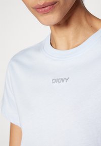 Šviesiai mėlynas medvilninis marškinėlis trumpomis rankovėmis ir apvaliu iškirpimu. Pagamintas su subtiliu rhinestone "DKNY" logotipu ant krūtinės. Lygus paviršius.