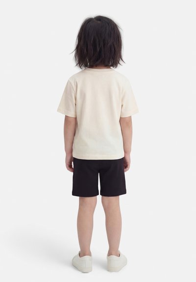 Enfant aux cheveux bruns mi-longs vu de dos, portant un t-shirt beige, un short noir et des baskets blanches sur fond blanc.