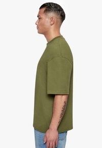 Olivegrünes, übergroßes T-Shirt, kurze Ärmel, Rundhalsausschnitt, aus Baumwolle, glatte Textur, getragen mit hellblauen Jeans, sichtbares Tattoo am Arm.