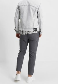Personne portant une veste en denim gris clair, une chemise à motifs, un pantalon gris foncé et des baskets blanches, se tenant de dos sur un fond uni.