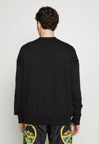 Černá mikina s kulatým výstřihem a dlouhými rukávy, na zadní straně s čtvercovou značkou. Materiál působí měkce a má texturu.